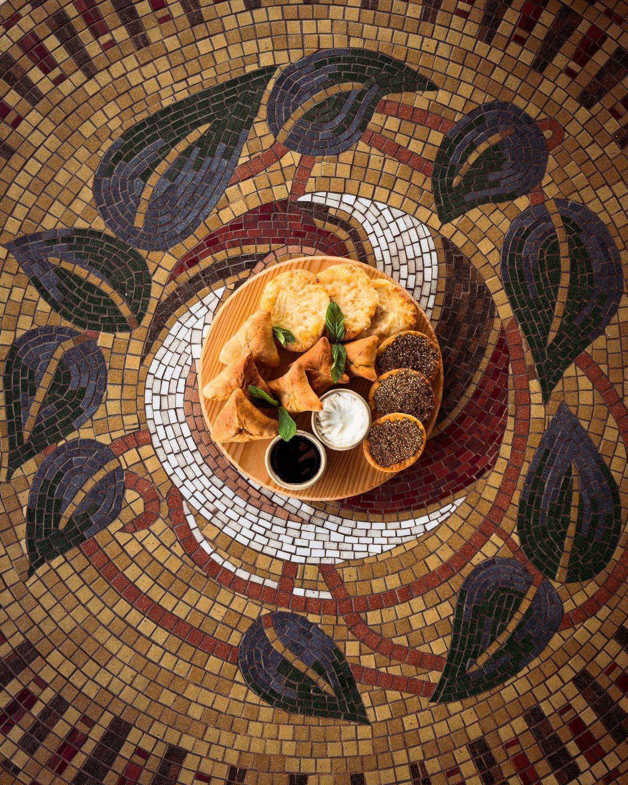 A round plate of baked bites with dips, on a leaf-pattern mosaic table seen from above